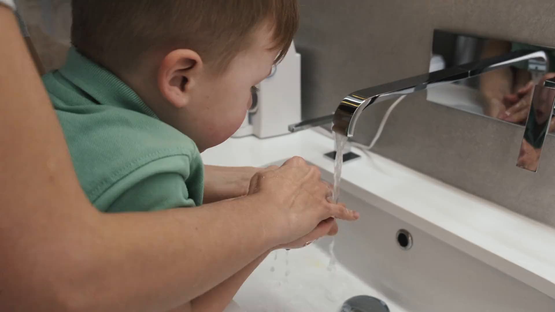 A parent's hands guiding a small child's hands under running water at a bathroom sink.